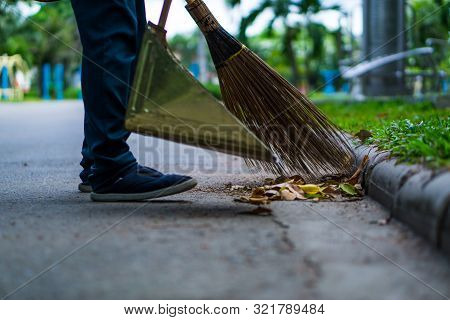 An Asian Woman Is Sweeping Dry Leaves By The Road In The Outdoor Garden. Cleaning Concept.
