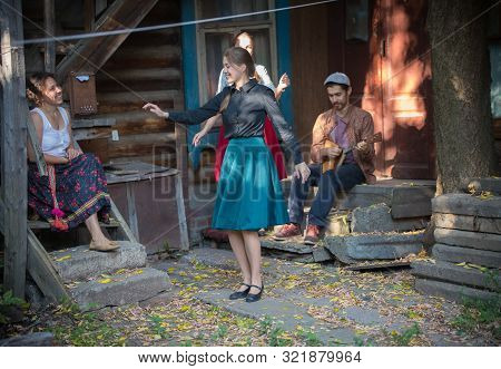A Pretty Woman In Blue Skirt Dancing By The Music Near The Country House - A Man Playing Balalaika