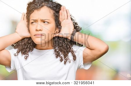 Young beautiful woman with curly hair wearing white t-shirt Trying to hear both hands on ear gesture, curious for gossip. Hearing problem, deaf