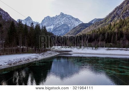 Beautiful View At Sunset Of The Peaceful Lake Fucine Tarvisio, Italy With Green Forest And Snowy Mou