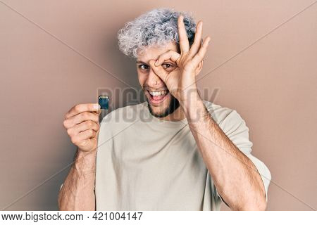 Young hispanic man with modern dyed hair holding sdxc card smiling happy doing ok sign with hand on eye looking through fingers 