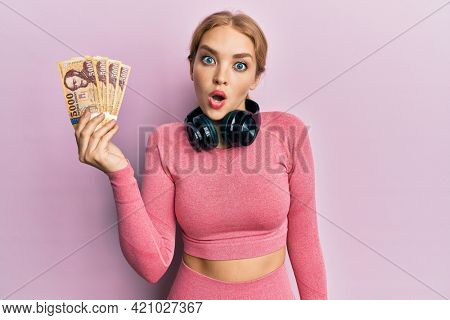 Beautiful blonde caucasian woman wearing gym clothes holding hungarian forints scared and amazed with open mouth for surprise, disbelief face 