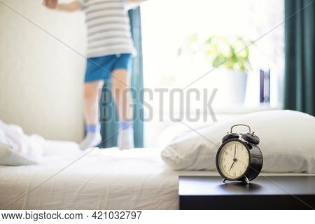 Just A Woken Up Little Child Jumping On The Bed With An Alarm Clock Standing On The Bedside Table In