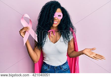 Middle age african american woman wearing super hero costume holding pink cancer ribbon celebrating achievement with happy smile and winner expression with raised hand 