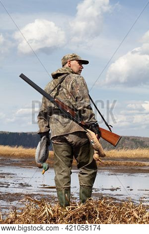 A Duck Hunter With A Duck Decoys In His Hands Stands On The Shallow Water. He's Got A Shotgun On His