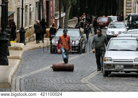 Azerbaijan, Baku - Nowember 2009: A Uniformed Worker Rolls A Gas Cylinder With His Foot Along The Ro