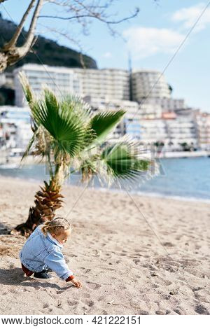 Little Girl In A Dress And Denim Jacket Sits On The Sand Against A Background Of Palm Trees On The B