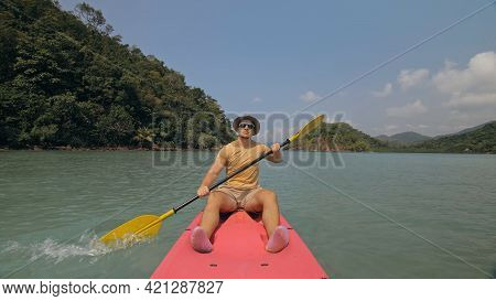 Man With Sunglasses And Hat Rows Pink Plastic Canoe Along Sea Against Green Hilly Islands With Wild 