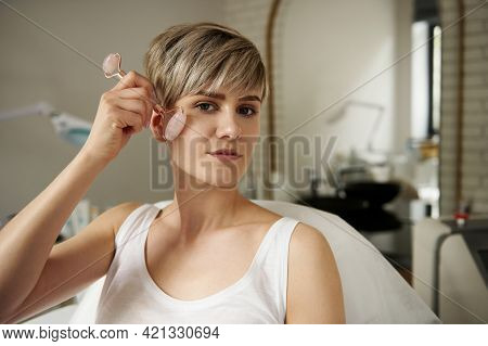 Attractive Blonde Woman Massaging Her Face With Jade Roller Sitting On A Massage Table In Cosmetolog