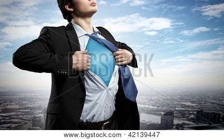 Image of young businessman showing superhero suit underneath his shirt standing against city background