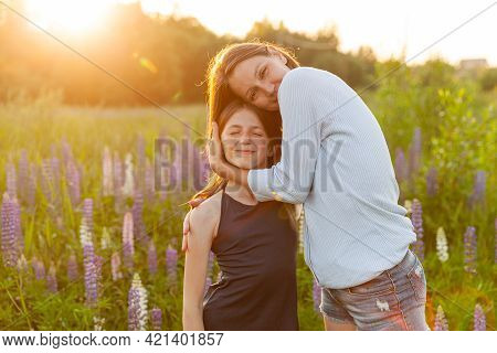 Young Mother Embracing Her Child Outdoor. Woman And Teenage Girl On Summer Field With Blooming Wild 