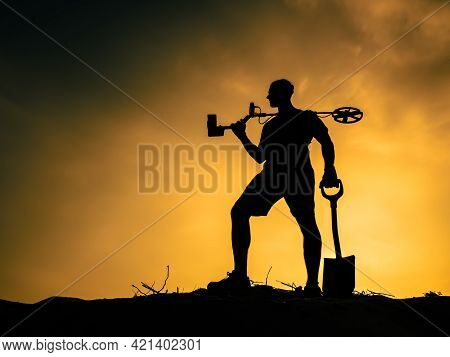Silhouette Of A Man Against The Sunset With A Metal Detector And A Shovel.