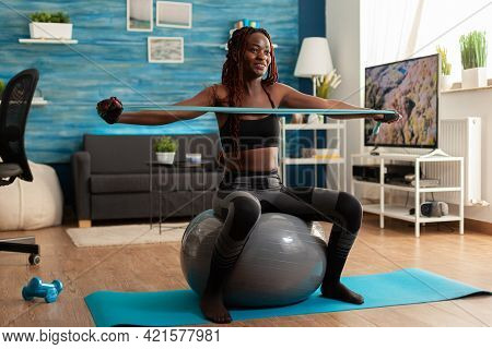 Athletic Fit Joyful Black Woman Working Out Back Muscle With Rubber Band, Sitting On Stability Ball 