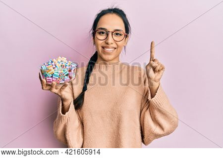 Young hispanic girl holding bowl of sugar candy smiling with an idea or question pointing finger with happy face, number one 