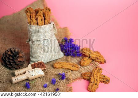 Borwn Cookie Stick Shape In Burlap Bag And Other With Flour Cocoa Dried Flower On Pink Background