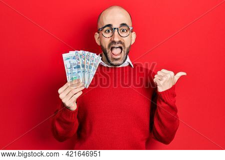 Young bald man holding peruvian sol banknotes pointing thumb up to the side smiling happy with open mouth 