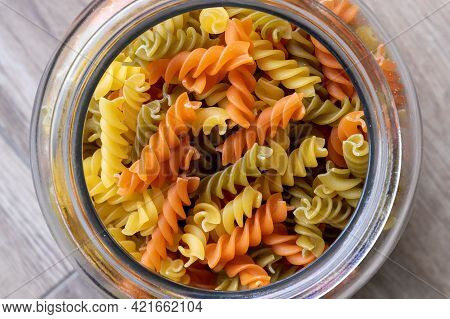 A Portrait Of A Glass Pot, Jar Or Bowl Full Of Spirelli In Three Different Colors. The Uncooked Tric