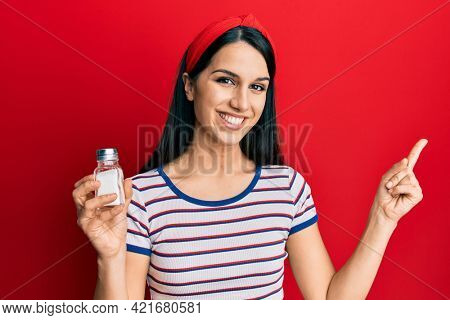 Young hispanic woman holding salt shaker smiling happy pointing with hand and finger to the side 