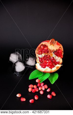 Broken Ripe Pomegranate Fruit With Leaves, Ice Cubes And Seeds On A Black Background.