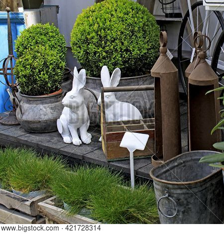 Two Ceramic Hares Among Boxwood Bushes And Garden Grass. Trolley On Wheels And Miscellaneous Gardeni