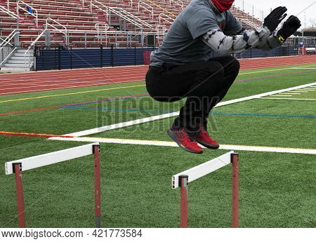A Male High School Track Runner Is Jumping Over Hurdles On A Green Turf Field During Track And Field