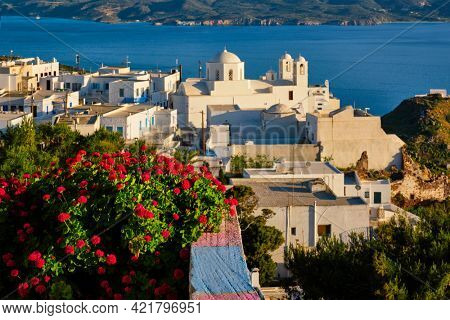 Picturesque scenic view of Greek town Plaka on Milos island over red geranium flowers and Orthodox greek church. Plaka village, Milos island, Greece. Focus on flowers