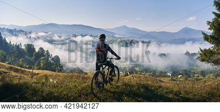 Man Riding Bicycle On Grassy Hill And Looking At Beautiful Misty Mountains. Male Bicyclist Enjoying 