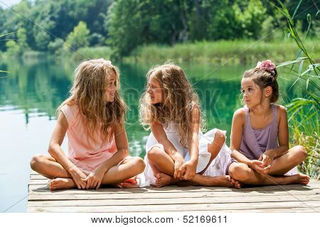 Three Girl Friends Sitting On Jetty At Lake.