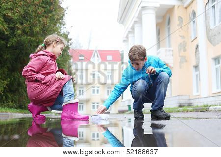 Boy and girl playing with paper boats in puddle on a cloudy day