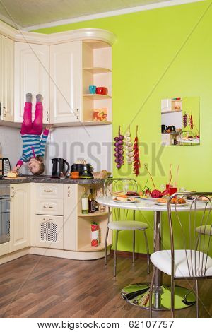 Little girl upside down sitting on the shelves in the kitchen with table and dishes
