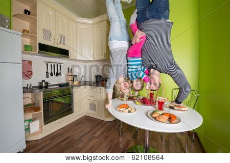 Family of three upside down holds a table in the kitchen with fridge and dishes