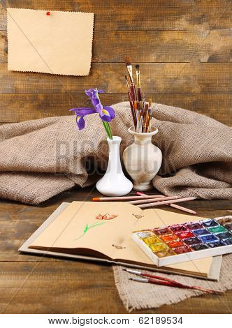 Composition with colorful watercolors, brushes in vase and sketcher on wooden background 