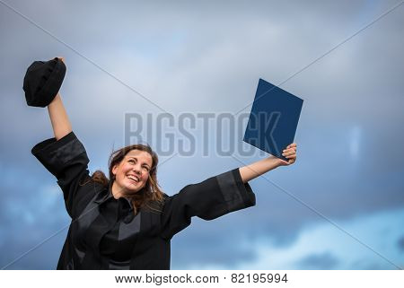 Pretty, young woman celebrating joyfully her graduation - spreading wide her arms, holding her diploma, savouring her success (color toned image; shallow DOF)