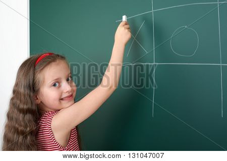 schoolgirl child in red striped dress drawing tic tac toe on green chalkboard background, summer school vacation concept