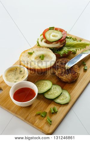 Cooking process of a sandwich burger ingredients on wooden cutting board on wooden table against white background fresh vegetables herbs fried meat buns sauces and knife shallow DOF