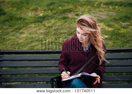Thoughtful attractive young woman sitting on bench and writing in notebook outdoors