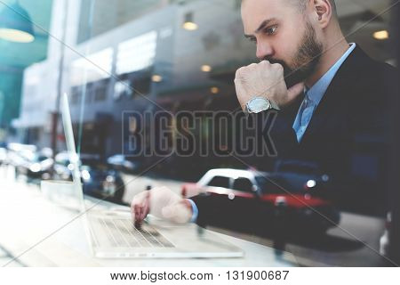 View through the window of confident pensive businessman is monitoring the shares on the stock market via portable net-book while is sitting in urban coffee shop of New York city. Double exposition