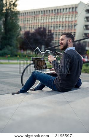 Young hipster enjoying coffee break outdoors while working on a computer.