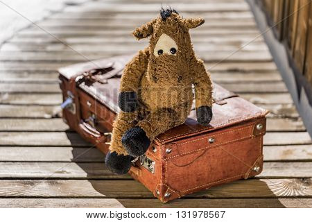 Single cute toy horse sitting in relaxed manner on suitcase over wooden deck outdoors