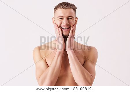 Portrait of young naked man applying aftershave and smiling while standing isolated on white background. Bearded hispter man standing shirtless and touching his face with hands. Men's skin care