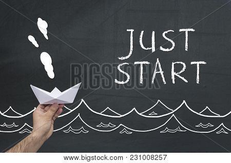 New Business Concept. Human Hand Holding Paper Boat Against Black Chalkboard With Text: Just Start.