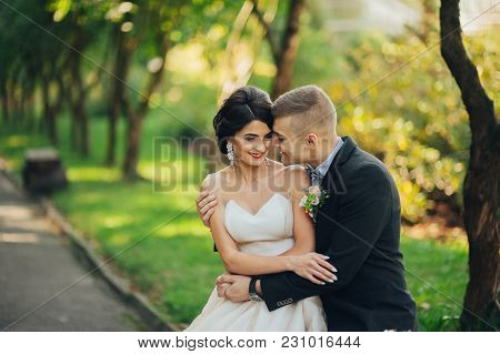 Beautiful Gorgeous Bride And Groom Walking In Sunny Park And Kissing. Happy Wedding Couple Hugging I