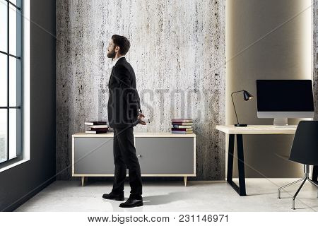 Side View Of Young European Businessman Looking Out Of Window In Modern Interior With Workpace And C