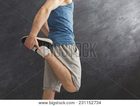 Young Man Warming Up Before Training At Gym. Unrecognizable Guy Stretching His Leg At Gray Backgroun