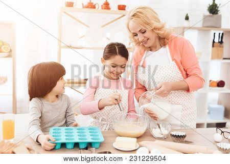 Small Cheerful Girl Is Beating Eggs In Kitchen. Grandmother Pours Milk Into Bowl. Grandson Keeps For