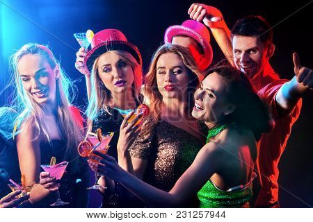 Dance party with group people dancing. Women and men have fun and drinking martini cocktail with citrus fruit in night club. Happy girl on foreground. Guys wearing hats. Rest after hard day at work.