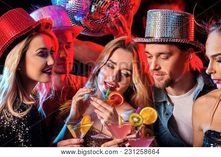 Dance party with group people dancing. Women and men have fun and drink martini cocktail with fruit in night club. Happy man in hat on foreground. Rest after hard day at work.