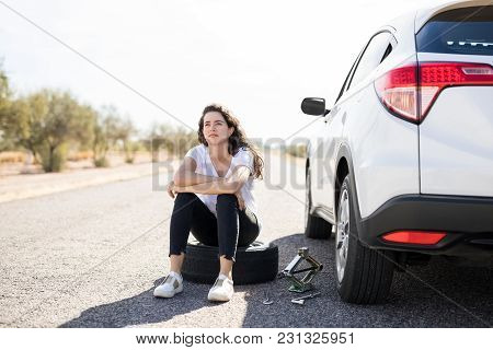 Portrait Of Upset Young Woman Sitting On Spare Tyre On Road While Changing The Flat Wheel Of Her Car