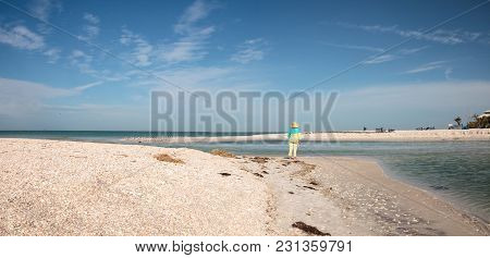 Older Woman Stands On The White Sand Beach In Front Of Aqua Blue Water Of Clam Pass