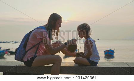 Travelers Mother And Little Daughter Sits At Seafront And Drinks Coconats In Trip. Mom Shares Her Co